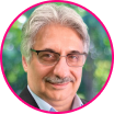 Portrait of an older man with gray hair, glasses, and a mustache, wearing a suit jacket, shown in a circular frame with a blurred outdoor background—an image that evokes trust and expertise in fraud prevention.